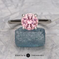 The "Anastasia" ring in 18k white gold features a 1.77-carat round pink tourmaline set above a rough blue stone on a textured surface, with "EARTH'S TREASURY" displayed below.