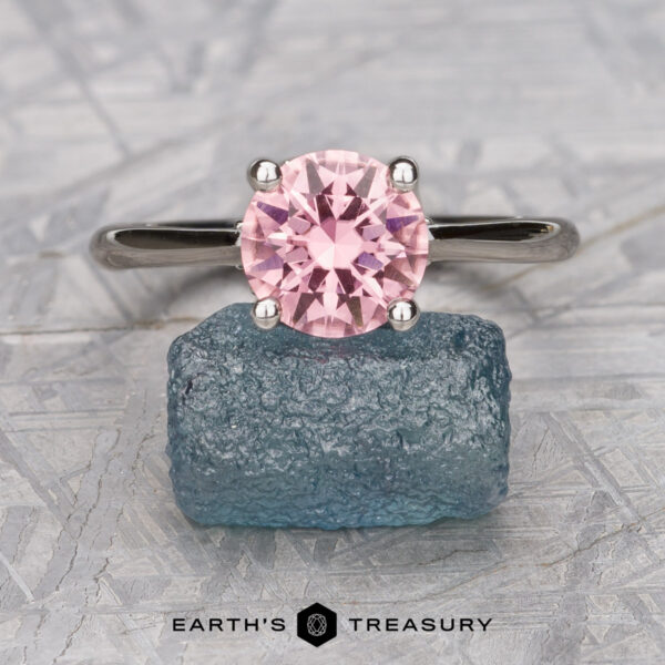 The "Anastasia" ring in 18k white gold with 1.77-Carat Tourmaline The "Anastasia" ring in 18k white gold features a 1.77-carat round pink tourmaline set above a rough blue stone on a textured surface, with "EARTH'S TREASURY" displayed below.