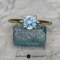 The "Alyssum" ring in 18K yellow gold with a 1.14-carat Montana sapphire is displayed upright on a rough blue stone, with “EARTH'S TREASURY” shown below.