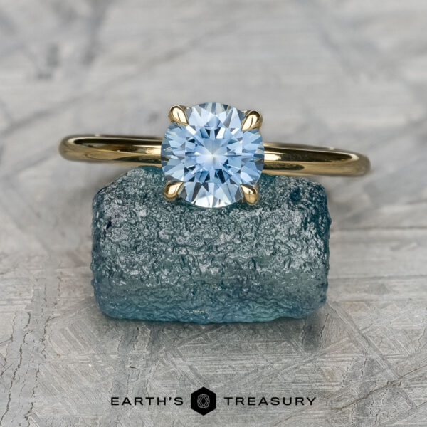 The "Alyssum" in 18K yellow gold with 1.14-carat Montana sapphire The "Alyssum" ring in 18K yellow gold with a 1.14-carat Montana sapphire is displayed upright on a rough blue stone, with “EARTH'S TREASURY” shown below.