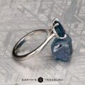 The "Anastasia" ring in platinum, featuring a 2.34-carat Montana sapphire in a Hidden Halo Solitaire style, is displayed upright against a rough blue stone on a textured metallic surface. "EARTH'S TREASURY" logo appears at the bottom.