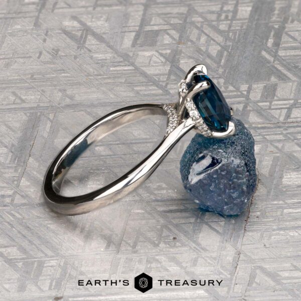 The "Anastasia" ring in platinum with 2.34-carat Montana sapphire The "Anastasia" ring in platinum, featuring a 2.34-carat Montana sapphire in a Hidden Halo Solitaire style, is displayed upright against a rough blue stone on a textured metallic surface. "EARTH'S TREASURY" logo appears at the bottom.