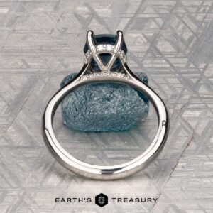 The "Anastasia" ring in platinum, featuring a 2.34-carat Montana sapphire and hidden halo diamonds, stands upright on a textured gray surface. The EARTH'S TREASURY logo and text are displayed at the bottom.
