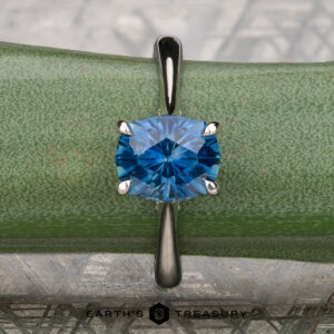 The Wetherell, Nikki and Yardy, Nicholas 5 ring features a blue gemstone in a four-prong silver Hidden Halo Solitaire setting, shown on a green textured surface with "Earth's Treasury" displayed at the bottom.