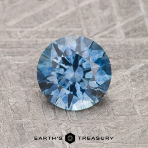 A 1.31-carat medium blue heated Montana Sapphire, cut in a round brilliant style, rests on a textured gray surface with "Earth's Treasury" at the bottom.