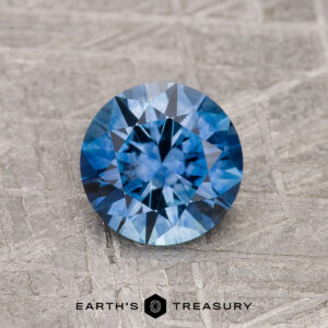 1.24-Carat Rich Blue Montana Sapphire (Heated) with a round brilliant cut displayed on textured gray, featuring the "EARTH'S TREASURY" logo and text below.