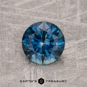 1.29-Carat Peacock Blue Montana Sapphire (Heated), round-cut, shown on a textured gray surface with the "Earth's Treasury" logo at the bottom.