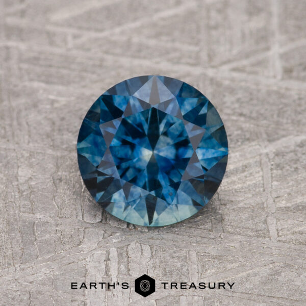 1.29-Carat Peacock Blue Montana Sapphire (Heated) 1.29-Carat Peacock Blue Montana Sapphire (Heated), round-cut, shown on a textured gray surface with the "Earth's Treasury" logo at the bottom.