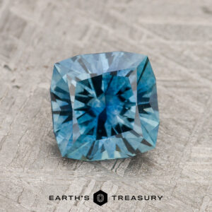 A 1.70-carat Teal Blue Montana Sapphire (Heated) with square cut and intricate facets rests on a textured gray surface. "EARTH'S TREASURY" is shown below the gemstone.