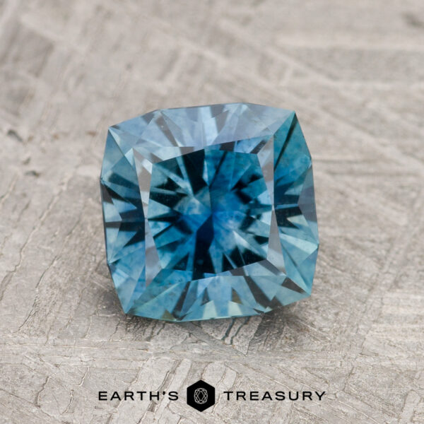 1.70-Carat Teal Blue Montana Sapphire (Heated) A 1.70-carat Teal Blue Montana Sapphire (Heated) with square cut and intricate facets rests on a textured gray surface. "EARTH'S TREASURY" is shown below the gemstone.