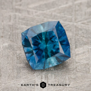 A 1.69-carat peacock blue Montana sapphire (heated), square-cut, is displayed on a textured surface with "Earth's Treasury" at the bottom.