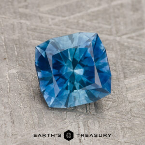 A 1.64-carat medium blue Montana sapphire (heated) with a square cushion cut rests on a textured gray surface, above the text "EARTH'S TREASURY" and a black hexagonal logo.