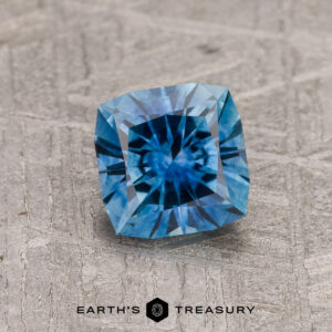 A 1.19-carat Peacock Blue Montana Sapphire (Heated), square-cut, is displayed on a textured gray surface featuring the "Earth's Treasury" logo at the bottom.