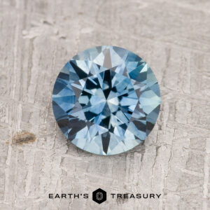 A 1.63-carat aqua blue Montana Sapphire (Heated), round brilliant-cut, is shown on a textured gray surface with "EARTH'S TREASURY" at the bottom.