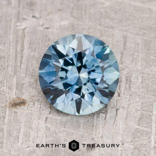1.63-Carat Aqua Blue Montana Sapphire (Heated) A 1.63-carat aqua blue Montana Sapphire (Heated), round brilliant-cut, is shown on a textured gray surface with "EARTH'S TREASURY" at the bottom.