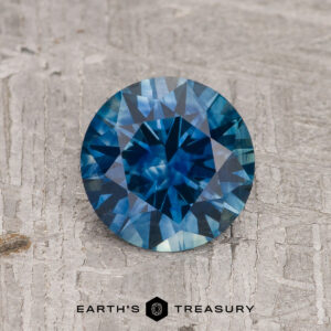 A 1.33-carat peacock blue Montana sapphire (heated) with a round brilliant cut rests on a textured gray wooden surface, with the "EARTH'S TREASURY" logo and text displayed below.