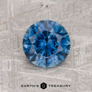 A 1.47-carat medium blue Montana Sapphire (heated), round and faceted, rests on a textured gray background with "EARTH'S TREASURY" below.