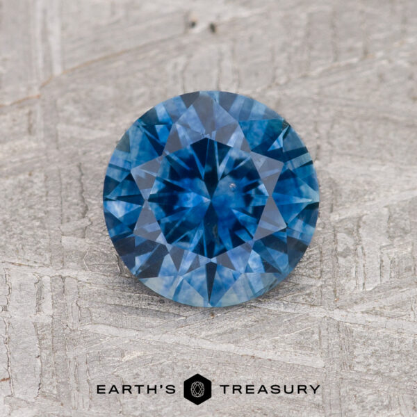 1.47-Carat Medium Blue Montana Sapphire (Heated) A 1.47-carat medium blue Montana Sapphire (heated), round and faceted, rests on a textured gray background with "EARTH'S TREASURY" below.
