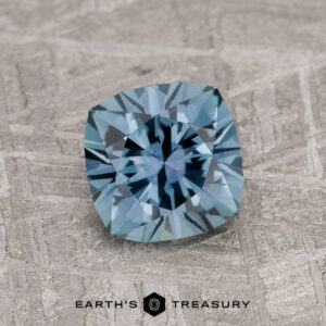 A 1.19-carat Steely Blue Montana Sapphire (Heated) with a cushion cut rests on a textured gray surface, featuring the "Earth's Treasury" logo and text at the bottom.