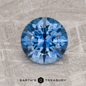 A 1.68-carat medium blue Montana Sapphire (heated), round and faceted, rests on a textured gray surface with the "Earth's Treasury" logo and text below it.