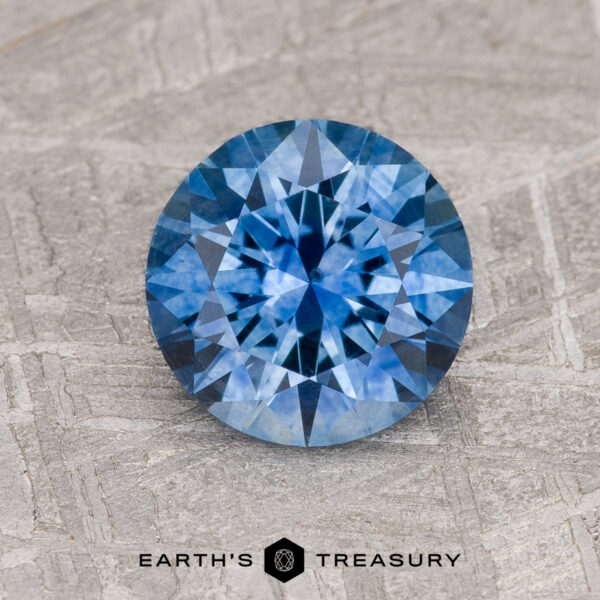 1.68-Carat Medium Blue Montana Sapphire (Heated) A 1.68-carat medium blue Montana Sapphire (heated), round and faceted, rests on a textured gray surface with the "Earth's Treasury" logo and text below it.