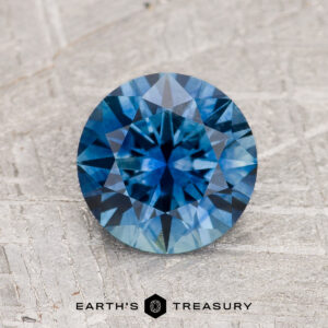 A 1.65-carat Medium Blue Montana Sapphire (Heated), round brilliant-cut, is displayed upright on a textured gray surface with the "Earth's Treasury" logo and text underneath.