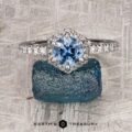 The “Ismene” ring in 14k white gold showcases a 1.29-carat Montana sapphire in a hexagonal halo with diamond accents, displayed on a rough blue stone with the Earth’s Treasury logo below.