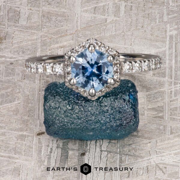 The “Ismene” ring in 14k white gold with 1.29-Carat Montana Sapphire The “Ismene” ring in 14k white gold showcases a 1.29-carat Montana sapphire in a hexagonal halo with diamond accents, displayed on a rough blue stone with the Earth’s Treasury logo below.