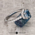 The “Ismene” ring in 14k white gold with a 1.29-carat Montana sapphire and petite pave diamond band rests on a rough blue stone atop a textured gray surface, with the “Earth’s Treasury” logo below.