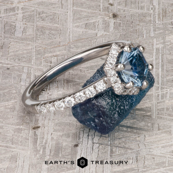 The “Ismene” ring in 14k white gold with 1.29-Carat Montana Sapphire The “Ismene” ring in 14k white gold with a 1.29-carat Montana sapphire and petite pave diamond band rests on a rough blue stone atop a textured gray surface, with the “Earth’s Treasury” logo below.