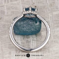 The “Ismene” ring in 14k white gold with a 1.29-carat Montana sapphire is displayed upright against a rough blue crystal on a textured surface, featuring the "Earth's Treasury" logo at the bottom.