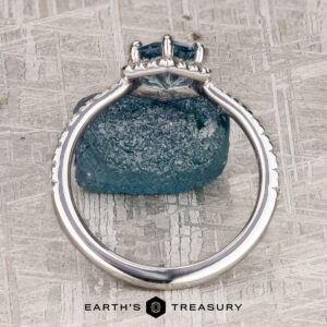 The “Ismene” ring in 14k white gold with a 1.29-carat Montana sapphire is displayed upright against a rough blue crystal on a textured surface, featuring the "Earth's Treasury" logo at the bottom.