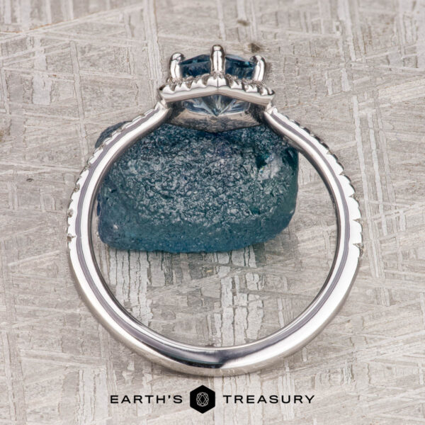 The “Ismene” ring in 14k white gold with 1.29-Carat Montana Sapphire The “Ismene” ring in 14k white gold with a 1.29-carat Montana sapphire is displayed upright against a rough blue crystal on a textured surface, featuring the "Earth's Treasury" logo at the bottom.