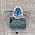 The "Pleione" ring in 14K white gold features a 1.31-carat pear-shaped Montana sapphire with a diamond halo and tapered pave band, displayed upright on a rough blue stone against a textured metallic background.