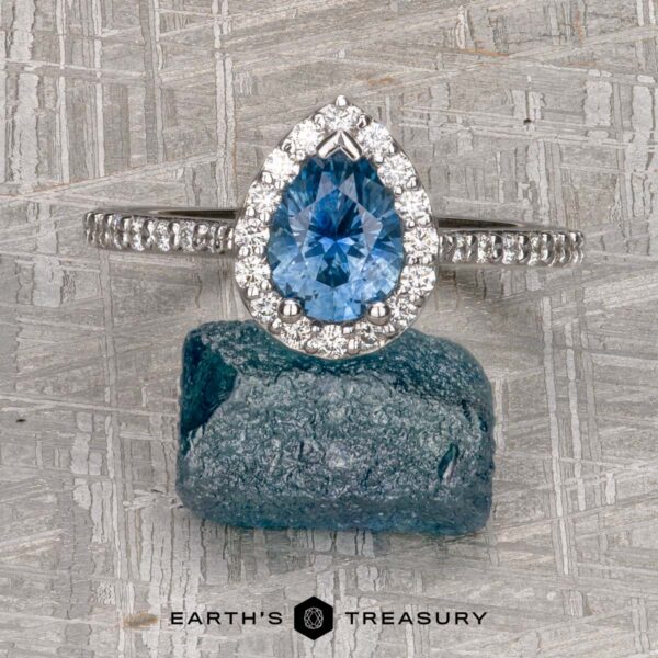 The "Pleione" ring in 14K white gold with 1.31-carat Montana sapphire The "Pleione" ring in 14K white gold features a 1.31-carat pear-shaped Montana sapphire with a diamond halo and tapered pave band, displayed upright on a rough blue stone against a textured metallic background.