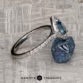 The "Pleione" ring in 14K white gold, featuring a 1.31-carat Montana sapphire and accent diamonds, rests against a rough blue stone on a textured surface. The text "EARTH'S TREASURY" appears at the bottom.