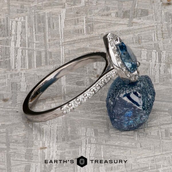 The "Pleione" ring in 14K white gold with 1.31-carat Montana sapphire The "Pleione" ring in 14K white gold, featuring a 1.31-carat Montana sapphire and accent diamonds, rests against a rough blue stone on a textured surface. The text "EARTH'S TREASURY" appears at the bottom.