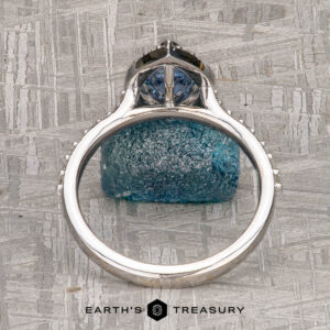 The "Pleione" ring in 14K white gold with a 1.31-carat Montana sapphire is displayed upright on a textured gray surface, featuring the Earth's Treasury logo at the bottom.