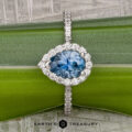 The "Pleione" ring in 14K white gold features a 1.31-carat pear-shaped Montana sapphire with a diamond halo and tapered pavé, shown on a green background with “Earth’s Treasury” text below.