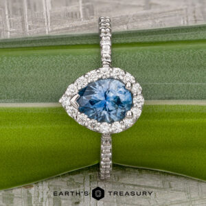 The "Pleione" ring in 14K white gold features a 1.31-carat pear-shaped Montana sapphire with a diamond halo and tapered pavé, shown on a green background with “Earth’s Treasury” text below.