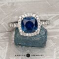 The Classic Pave Halo Ring in Platinum features a 1.88-carat Montana sapphire center with sparkling clear accents, elegantly displayed on a rugged blue mineral backdrop.