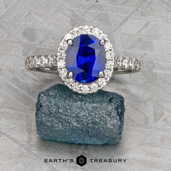 The Classic Pave Halo Ring in 14k white gold with 1.96-carat Madagascar sapphire The Classic Pave Halo Ring in 14k white gold, featuring a 1.96-carat Madagascar sapphire surrounded by pavé stones, is displayed on rough blue mineral with "Earth's Treasury" text below.