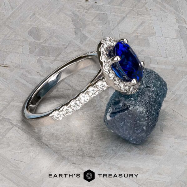 The Classic Pave Halo Ring in 14k white gold with 1.96-carat Madagascar sapphire The Classic Pave Halo Ring in 14k white gold, showcasing a 1.96-carat Madagascar sapphire and diamond accents, rests on a rough blue stone, with the Earth’s Treasury logo displayed below.