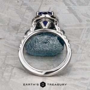 The Classic Pave Halo Ring in 14k white gold with a 1.96-carat Madagascar sapphire is displayed upright above a matching blue stone on a textured gray surface, with the "Earth's Treasury" logo at the bottom.