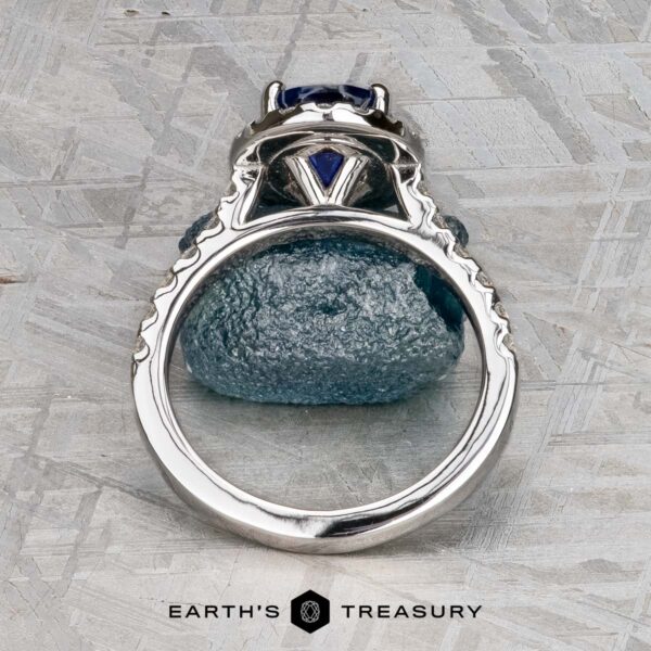 The Classic Pave Halo Ring in 14k white gold with 1.96-carat Madagascar sapphire The Classic Pave Halo Ring in 14k white gold with a 1.96-carat Madagascar sapphire is displayed upright above a matching blue stone on a textured gray surface, with the "Earth's Treasury" logo at the bottom.