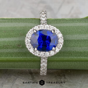 The Classic Pave Halo Ring in 14k white gold features a 1.96-carat Madagascar sapphire center, surrounded by sparkling white stones, displayed on a green-striped surface.