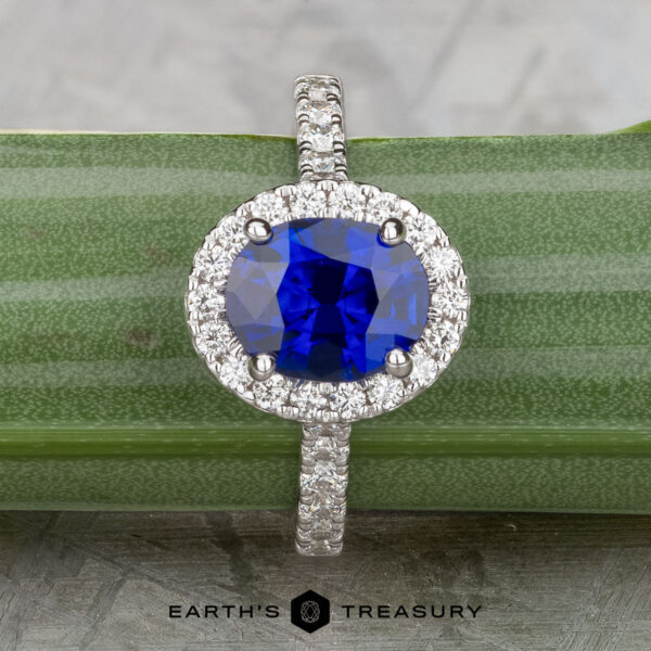 The Classic Pave Halo Ring in 14k white gold features a 1.96-carat Madagascar sapphire center, surrounded by sparkling white stones, displayed on a green-striped surface.