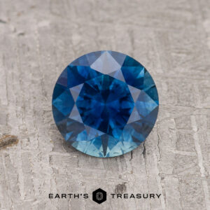 The 1.32-Carat Rich Peacock Blue Montana Sapphire (Heated), round brilliant-cut, is displayed on a textured gray surface with the "Earth's Treasury" logo and text beneath the stone.
