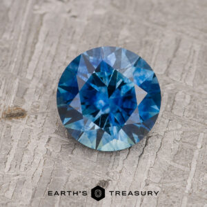 A 1.40-carat medium blue Montana Sapphire (heated), round and faceted, rests on a textured gray surface above the “EARTH’S TREASURY” text and a geometric logo.