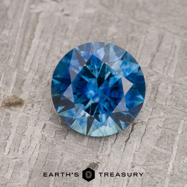 1.40-Carat Medium Blue Montana Sapphire (Heated) A 1.40-carat medium blue Montana Sapphire (heated), round and faceted, rests on a textured gray surface above the “EARTH’S TREASURY” text and a geometric logo.